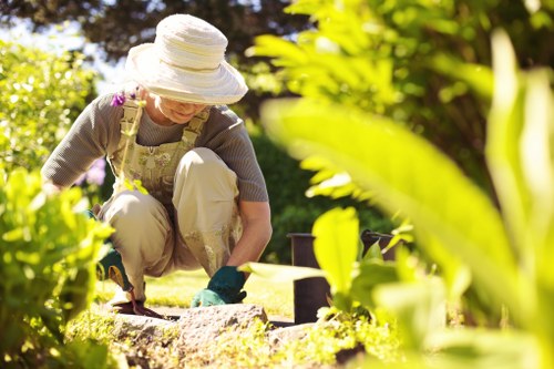 A woman wearing a wide-brimmed hat, beige trousers, and gardening gloves is tending to a garden, crouched down close to the ground while working on soil or plants. The garden features a lush, green lawn in the foreground, with various shrubbery and leafy plants framing the scene. In the background, there are flowering bushes or small trees that add natural colour and texture to the outdoor space. The sunlight illuminates the scene, indicating a clear day, and the environment appears well-maintained and vibrant. This setting reflects typical gardening activities that local landscape garden services, such as those by Landscapers Canary Wharf, might assist with, including planting, soil preparation, and general outdoor maintenance, suitable for gardens in the Canary Wharf area near London postcode E14.