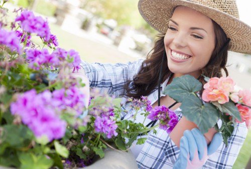 A woman wearing a straw hat and blue gardening gloves is smiling while tending to a variety of vibrant flowering plants in a well-maintained outdoor garden. The garden features a lush green lawn in the foreground, bordered by flower beds with purple and pink blooms, alongside leafy shrubs. The background shows a landscaped area with trees and a paved patio, indicating a thoughtfully designed outdoor space suitable for relaxation and outdoor activities. The scene is illuminated by natural sunlight, emphasizing the fresh, natural tones of the garden elements and suggesting a calm, pleasant weather. The woman's engagement with the plants highlights care and maintenance typical of professional garden renovation services provided by companies like Landscapers Canary Wharf, aligning with landscaping and gardening improvement themes for residential outdoor spaces in Canary Wharf or nearby London areas.