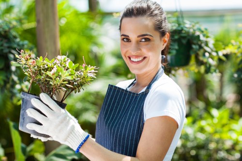 A young woman with dark hair tied back, smiling and wearing a striped apron and white gloves, is holding a small potted shrub with green and purple leaves in a garden setting. The background features lush greenery, including trees, bushes, and a wooden post, with a sunny outdoor environment indicative of a well-maintained garden or landscaped outdoor space typical of Canary Wharf. The scene reflects gardening activity, possibly related to planting or tending to garden elements, aligning with landscaping and lawn care services offered by Landscapers Canary Wharf. Natural light highlights the vibrant foliage and the woman's cheerful expression, emphasizing professional outdoor gardening work in a local London setting with a focus on garden maintenance and planting services.