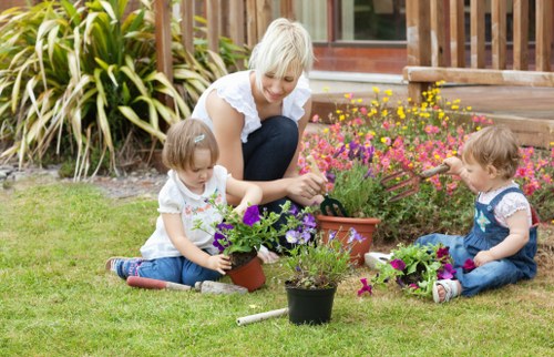 A woman with light blonde hair, wearing a white short-sleeved blouse and dark jeans, is sitting on a green lawn in a garden, engaging with two young children. The children, both girls with light brown hair, are seated on the grass, each tending to potted plants with purple and pink flowers. The garden features a manicured lawn, with flower beds containing various colorful blooms, a wooden deck with stairs, and dense foliage of ornamental grasses and shrubs in the background. The scene is illuminated by natural daylight, suggesting a clear weather day, and showcases a well-maintained outdoor space that highlights gardening activities typical for suburban or urban gardens near Canary Wharf, supporting local landscaping and eco-friendly practices promoted by Landscapers Canary Wharf.