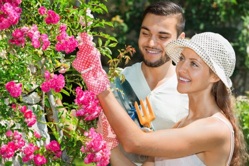 In a well-maintained garden scene, a young woman and a young man are engaged in gardening tasks; the woman is wearing a white sun hat, pink gardening gloves, and holding flowering pink roses, while the man, dressed casually, stands beside her, smiling. The garden features a lush, green environment with dense foliage, a variety of shrubs, and neatly trimmed hedges in the background. Bright pink roses are in full bloom along a trellis or garden support, contrasting with the green leaves and soil. A wooden or paved patio area is partially visible in the foreground, indicating a landscaped outdoor space designed for leisure and gardening. The lighting suggests a clear, sunny day, emphasizing the vibrant colours and natural textures of the garden elements. This scene reflects typical features of residential gardens in Canary Wharf, including organized planting areas and outdoor maintenance, supporting professional gardening services offered by companies such as Landscapers Canary Wharf.