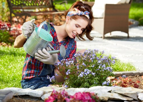 A young woman with light skin, wearing a colorful plaid shirt, gray gardening gloves, and a floral headband, is kneeling on a stone-edged garden border in a landscaped outdoor space in Canary Wharf. She is smiling as she waters a small flowering shrub with light purple blossoms using a metal watering can. Behind her, there is a paved patio area with outdoor furniture, including a chair, and lush green grass and trees in the background. The garden features a neatly maintained lawn, flower beds with various plants, and a mix of natural soil and stone borders, all under natural daylight conditions. The scene reflects a well-maintained outdoor area suitable for gardening and landscaping services, with a focus on flower bed care and outdoor plant maintenance typical of residential gardens in the area.