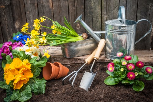 A garden scene featuring a variety of gardening tools and containers arranged on rich, dark soil. In the foreground, there are vibrant flowering plants including yellow daffodils and pink primroses with green foliage. Behind these, a small empty terracotta plant pot is positioned next to a metal watering can with a long spout. To the left, a rectangular metal container holds a bunch of yellow daffodils, while a garden fork and trowel with wooden handles are leaning against the soil. The background consists of a wooden fence that encloses the outdoor space, suggesting a typical landscaped garden or backyard. The scene depicts preparations for gardening or planting activities in a well-maintained outdoor area, characteristic of the gardening services offered by Landscapers Canary Wharf in London, with natural lighting indicating a clear, bright day.