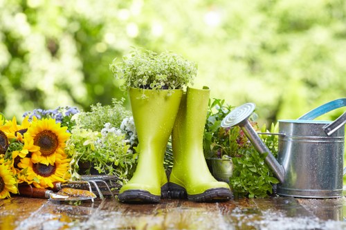 A garden scene featuring a wooden outdoor table with a pair of bright green rubber gardening boots placed as a vase holding delicate white and green flowering plants. To the left of the boots, there is a bouquet of vibrant yellow sunflowers and purple flowers, while on the right, a metal watering can with a detachable spout and a blue-handled garden trowel are positioned. The background shows a lush, green, blurred garden with dense foliage and trees, indicating a healthy outdoor environment. The surface of the table appears slightly damp, suggesting recent watering or rain, and the scene is illuminated by natural daylight, reflecting a typical garden setting in the Canary Wharf area. This image emphasizes gardening tools and plant care elements that are integral to professional landscaping and planting projects near urban green spaces like Canary Wharf, where curated gardens and flower beds are a common feature.
