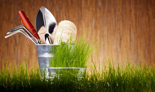 A close-up image of a small, outdoor garden scene featuring a patch of lush, green grass in the foreground. Positioned on the grass are two galvanized metal buckets containing gardening tools, including a small hand trowel, a hand fork, and a pair of pruning shears, along with a ball of natural twine. The background shows a warm-toned wooden surface, likely a garden shed or fence, creating a rustic atmosphere. The scene suggests a gardening or outdoor maintenance setting typical of residential gardens in Canary Wharf or surrounding areas, with natural lighting indicating daytime conditions. This setting reflects a focus on gardening activities such as planting, pruning, or general garden care, themes relevant to landscaping and outdoor maintenance services provided by Landscapers Canary Wharf.