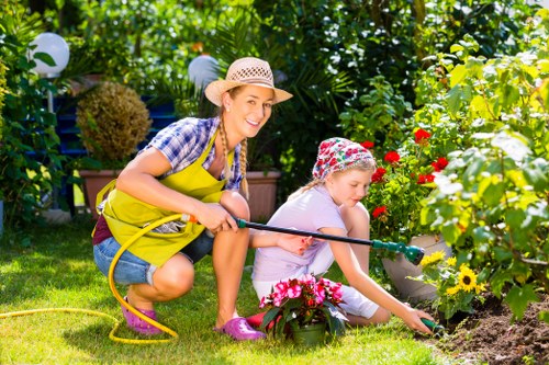 In a lush, well-maintained garden, two young girls are engaged in outdoor gardening activities. The older girl, wearing a straw hat, yellow apron, a blue checkered shirt, and pink gardening boots, is holding a garden hose and smiling at the camera, suggesting she is watering plants. The younger girl, dressed in a light grey outfit with a colorful floral hat, is focused on planting or tending to flower beds, which are filled with vibrant red, yellow, and pink blooms. The garden features a neatly trimmed lawn, bordered by flower beds and potted plants, with dense green foliage and trees providing a natural backdrop. String lights are visible hanging from the foliage, indicating careful garden landscaping and outdoor decoration typical of a residential outdoor space in London postcodes. The scene captures a sunny day with bright, natural lighting, emphasizing the health and vibrancy of the garden. Landscapers Canary Wharf offers gardening services that can help create similar inviting outdoor environments, suitable for families and outdoor leisure.