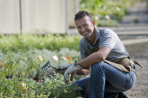 A man kneeling in a neatly maintained garden, surrounded by lush green plants and flowering bushes, holding a garden fork and smiling at the camera. The garden features a grass lawn in the foreground, bordered by a paved pathway and a wooden fence in the background, with mature trees providing natural shade. The scene is set outdoors under natural daylight with clear weather, illustrating typical gardening activities that might be offered by Landscapers Canary Wharf to enhance residential outdoor spaces in the local area.